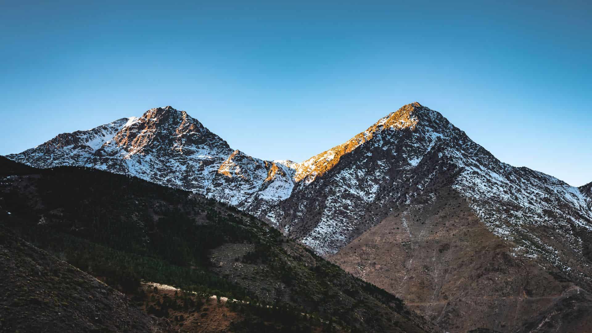 Two snow-capped mountain peaks under a clear blue sky, with sunlight hitting one summit.
