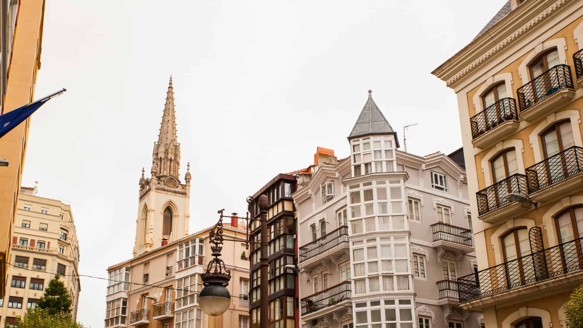 Colorful buildings and a church steeple rise against a cloudy sky in a European city street scene.