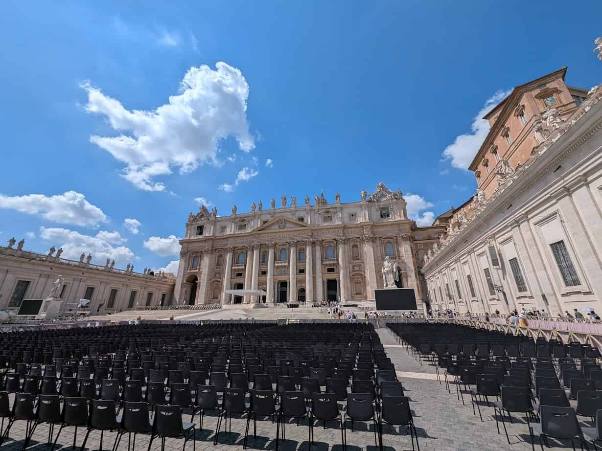 Rows of empty black chairs facing St. Peter’s Basilica in Vatican City under a bright blue sky with clouds—an iconic sight perfect for your Family Itinerary when exploring Rome with Kids.