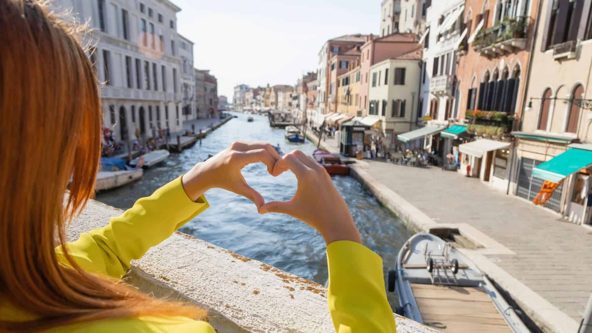 A person forms a heart shape with their hands overlooking a canal in a European city.