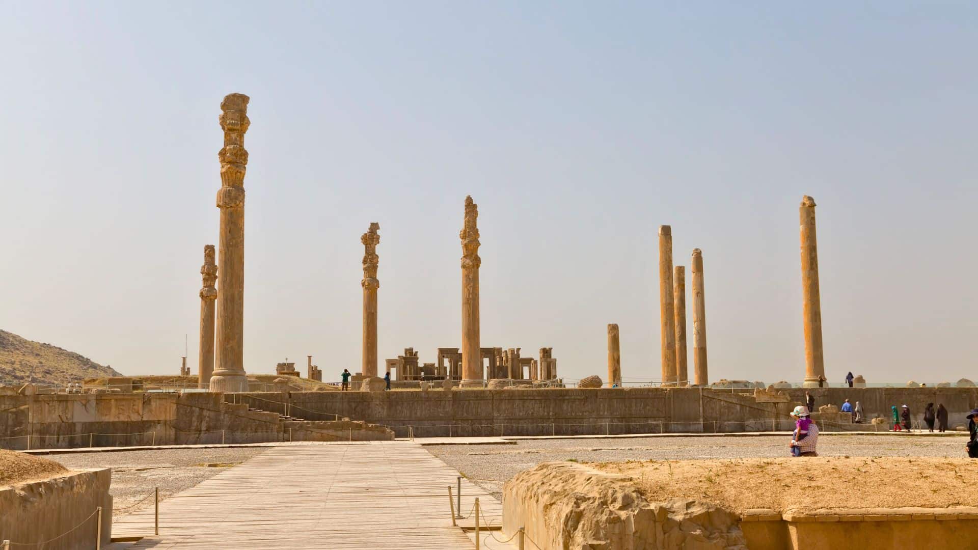 Ancient stone columns and ruins of Persepolis under a clear sky, with people walking around the site.