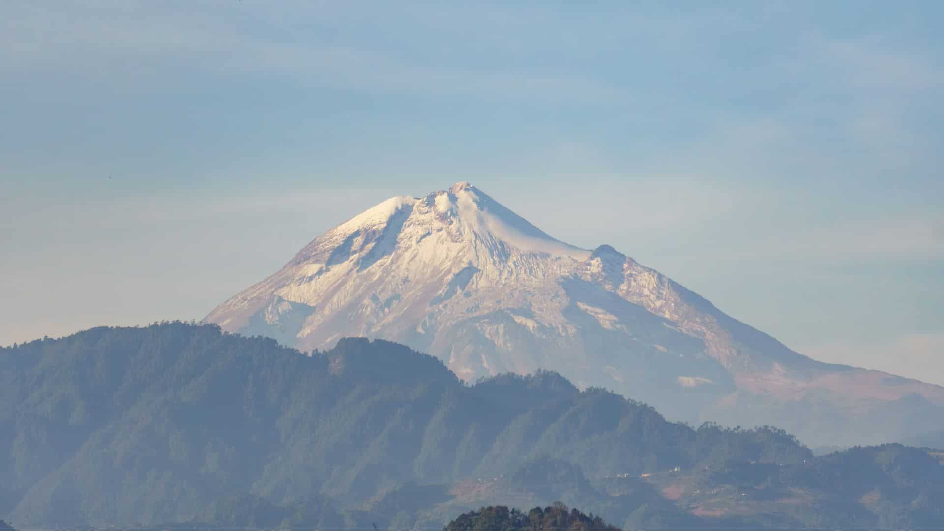Snow-capped volcano with green hills in the foreground under a clear blue sky.