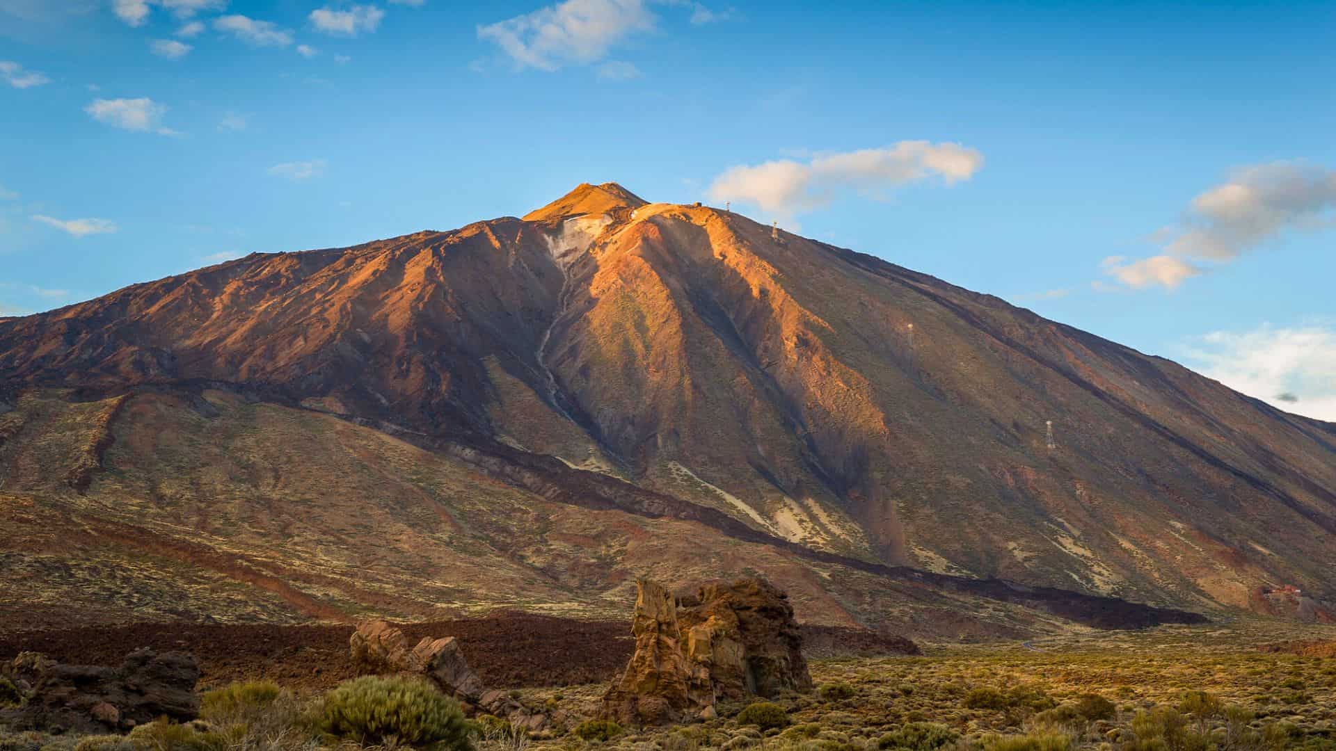 Mountain with rocky slopes and patches of greenery under a blue sky with scattered clouds at sunset.