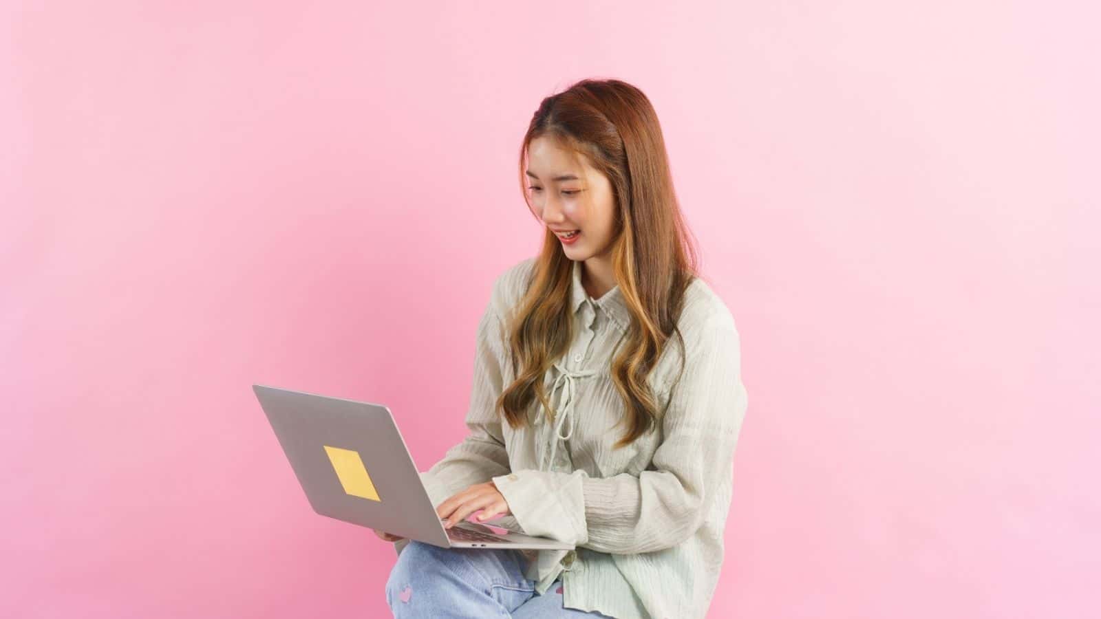 A young woman using a laptop, smiling, seated against a plain pink background.