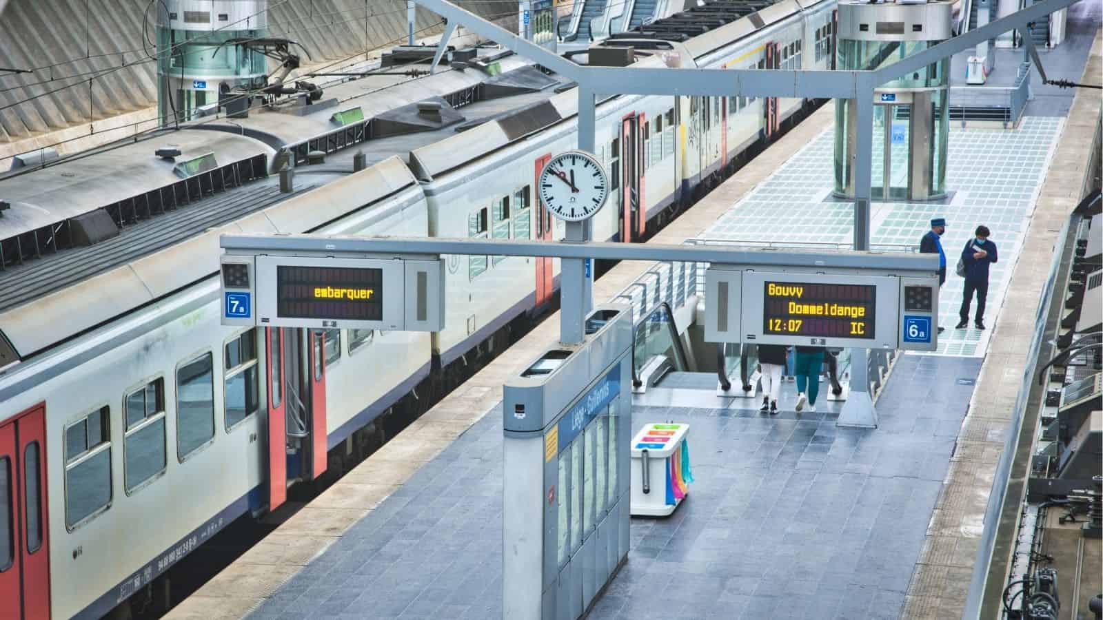 Train station platform with a train, clock, digital signs, and a few people waiting or walking.