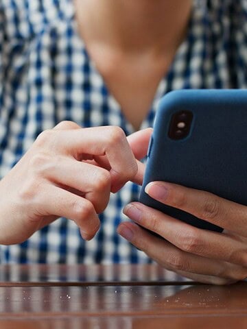 Person in a checkered shirt using a smartphone at a wooden table, with a smartwatch on their wrist.