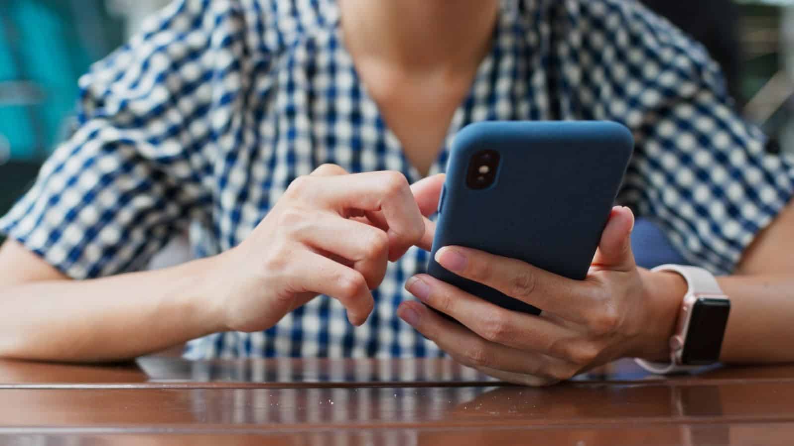 Person in a checkered shirt using a smartphone while sitting at a wooden table.