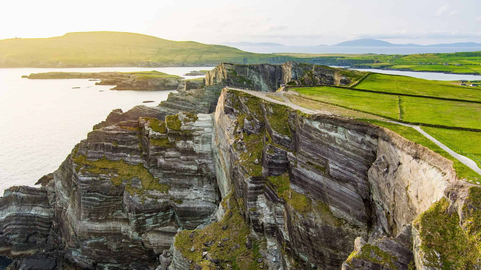 Steep, rocky sea cliffs with green fields above and water below under a bright, clear sky.