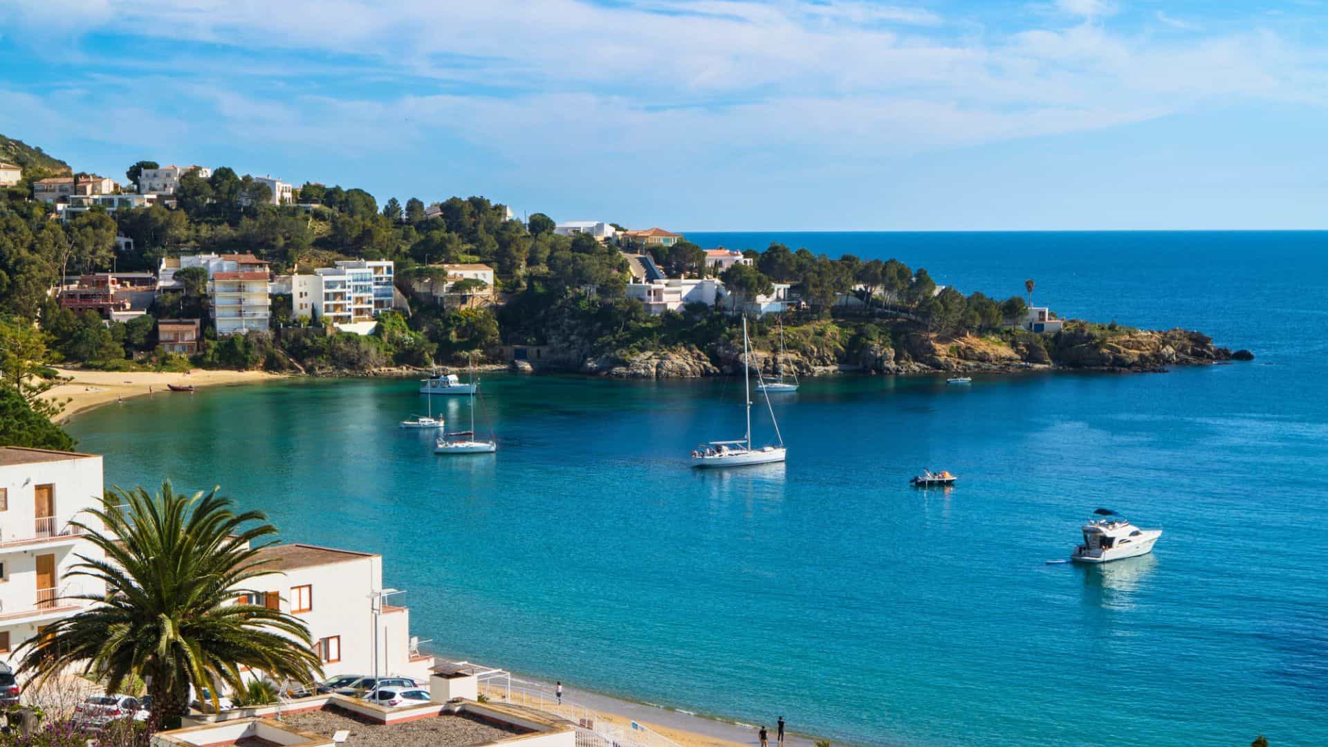 Boats anchored in a calm blue bay surrounded by houses, trees, and a sandy beach under a clear sky.