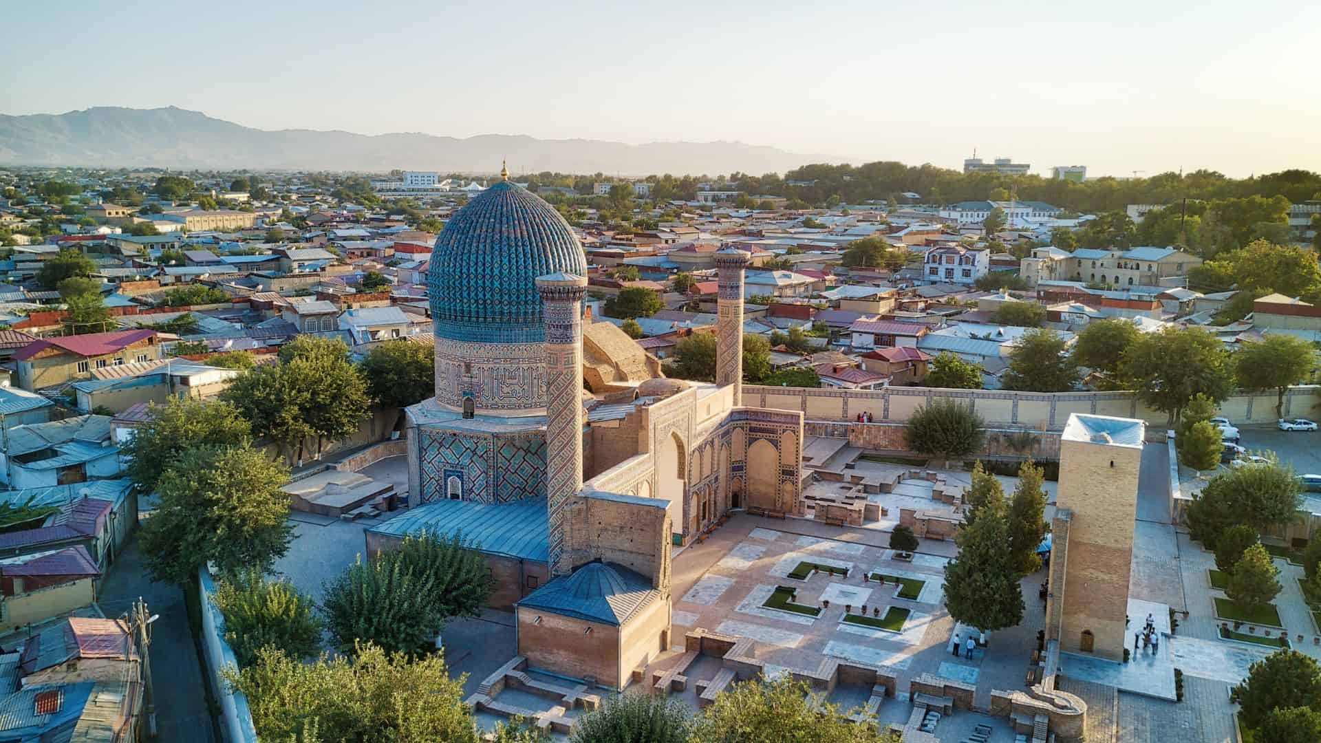 Aerial view of a blue-tiled domed mausoleum surrounded by a cityscape and mountains in the background.