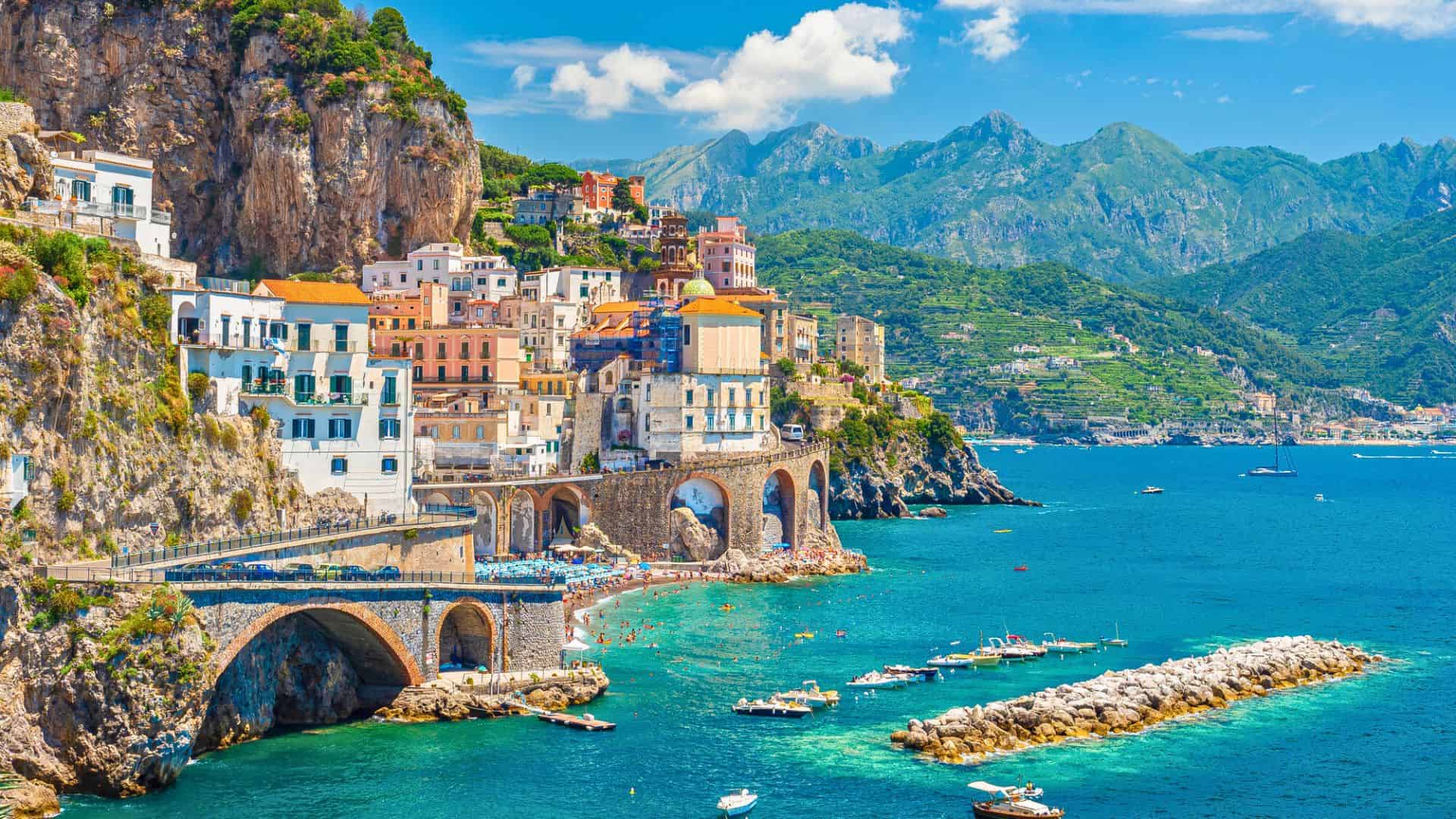 Colorful cliffside buildings overlook a blue sea with boats and a rocky pier, set against green mountains.