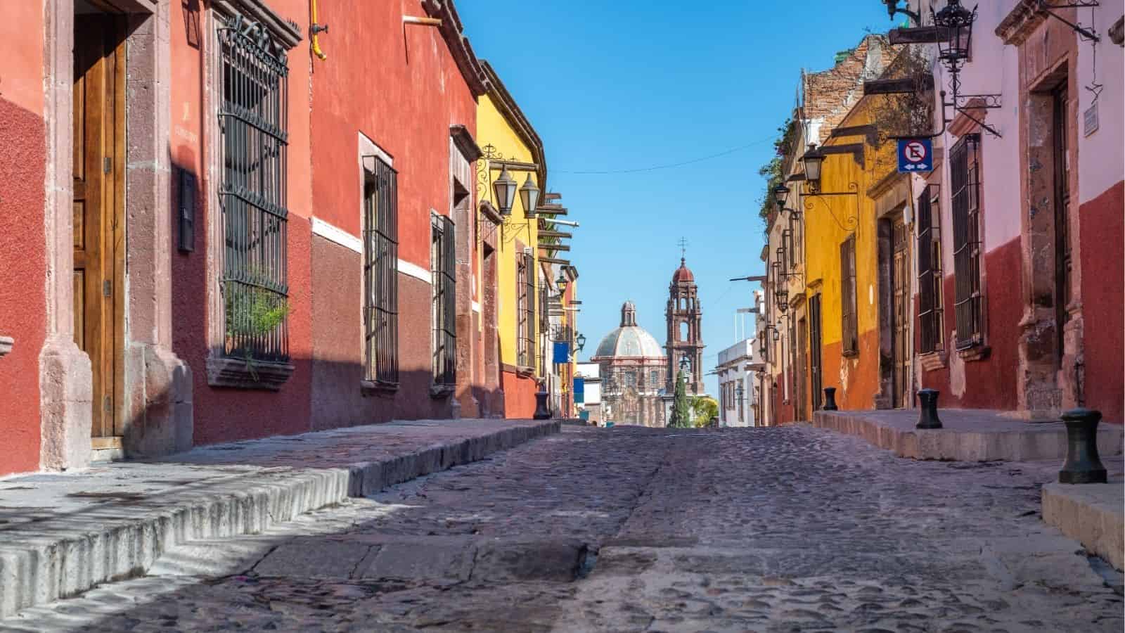 Colorful cobblestone street lined with colonial buildings, leading to a distant domed church under a blue sky.