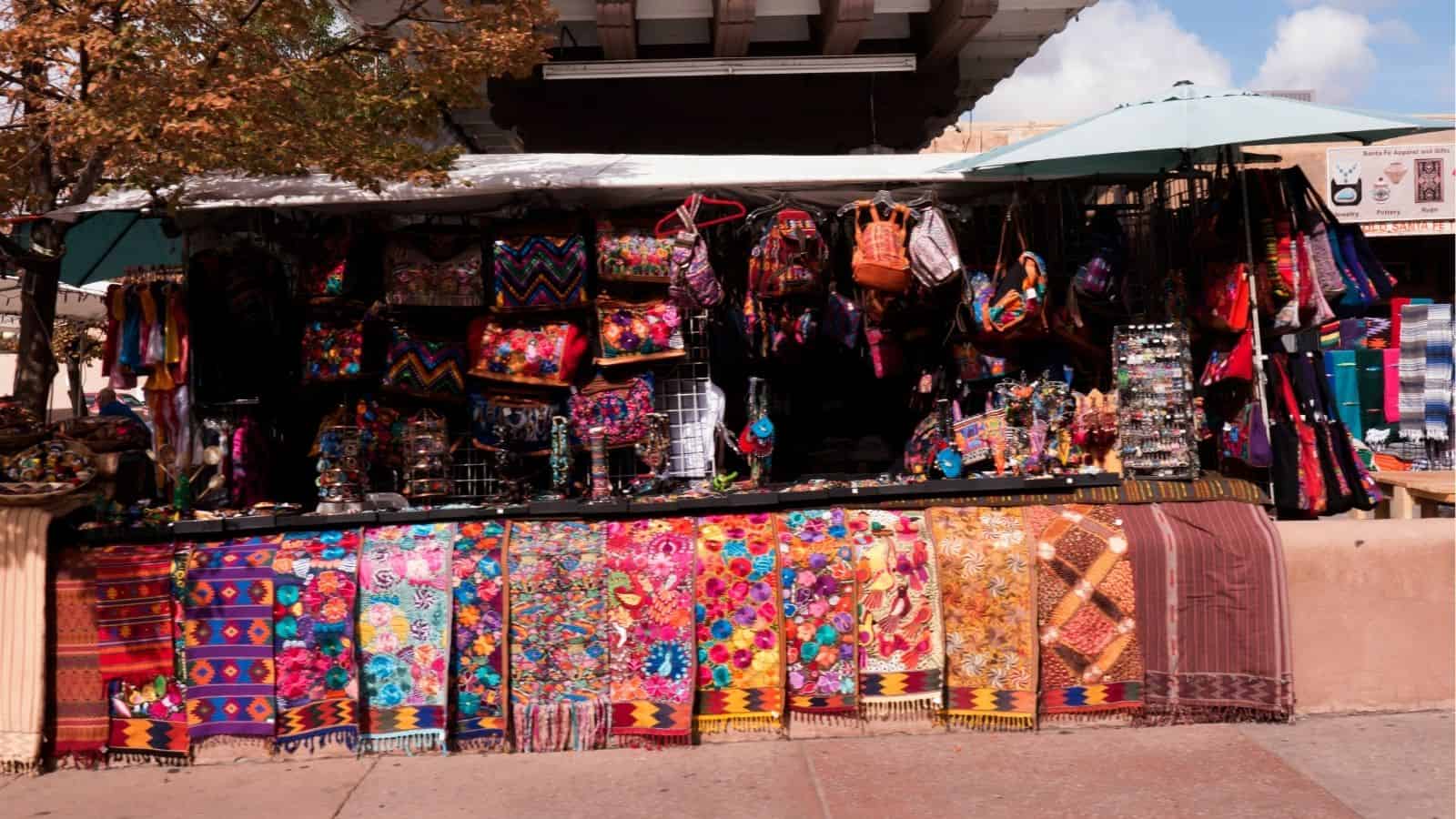 A market stall displays colorful woven textiles, bags, and handmade crafts under an umbrella.