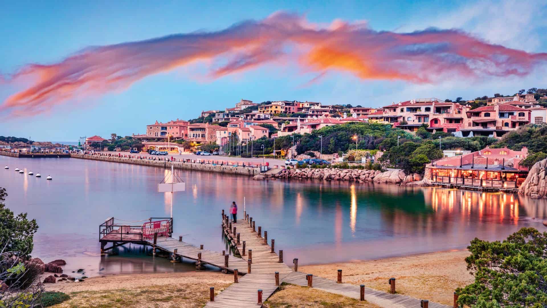 Colorful seaside village with pastel houses, wooden pier, and calm water under a vibrant, streaked sunset sky.