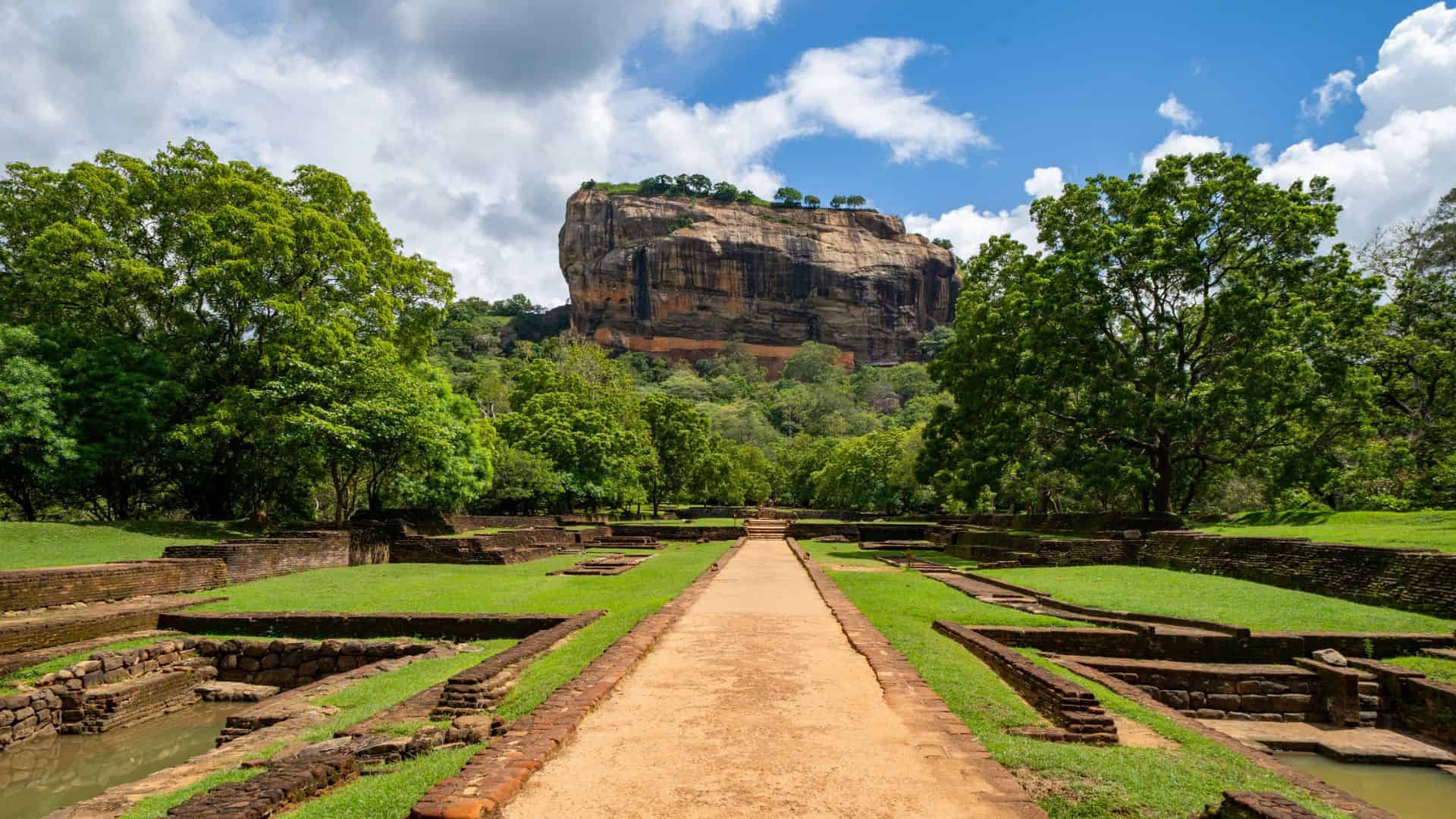 A stone path leads to Sigiriya Rock Fortress, surrounded by lush trees under a bright, partly cloudy sky.