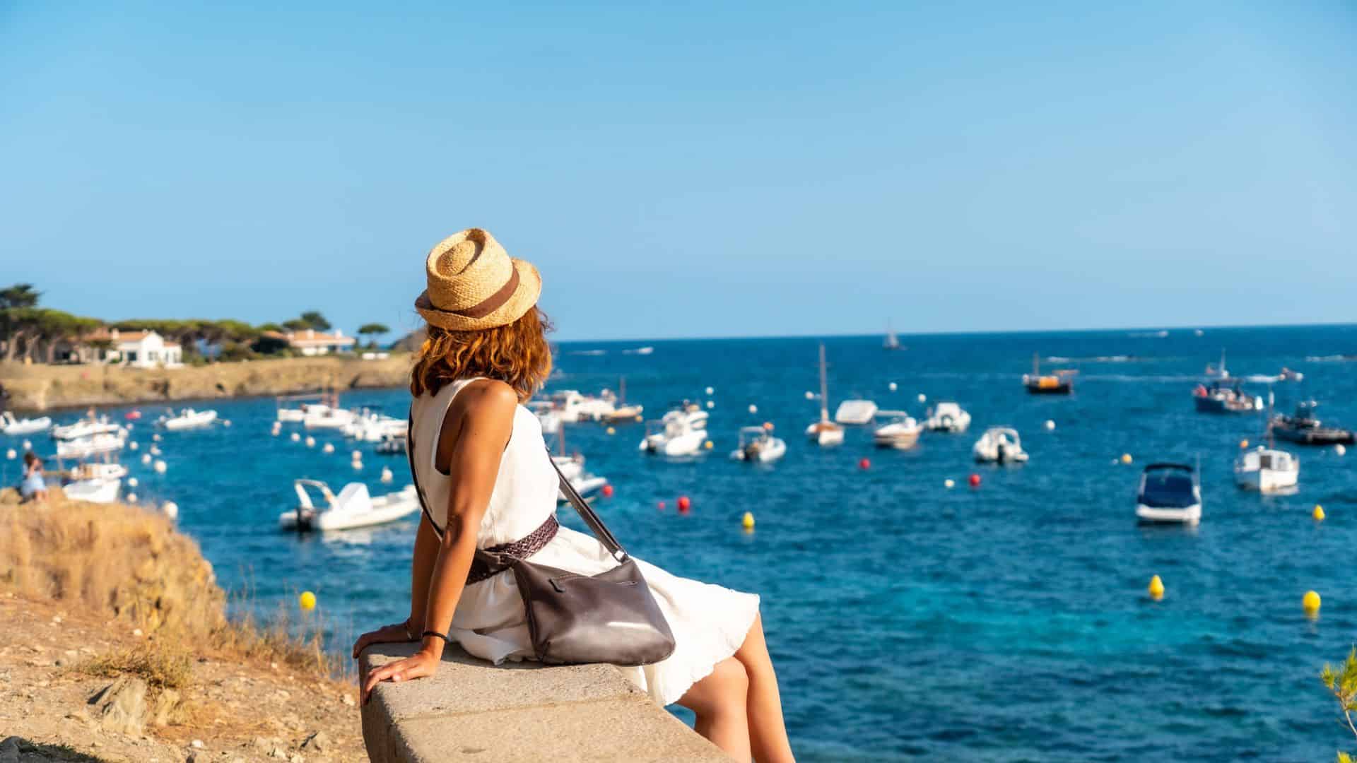 Woman in a sunhat sits on a wall overlooking a bay with boats on a bright, sunny day.