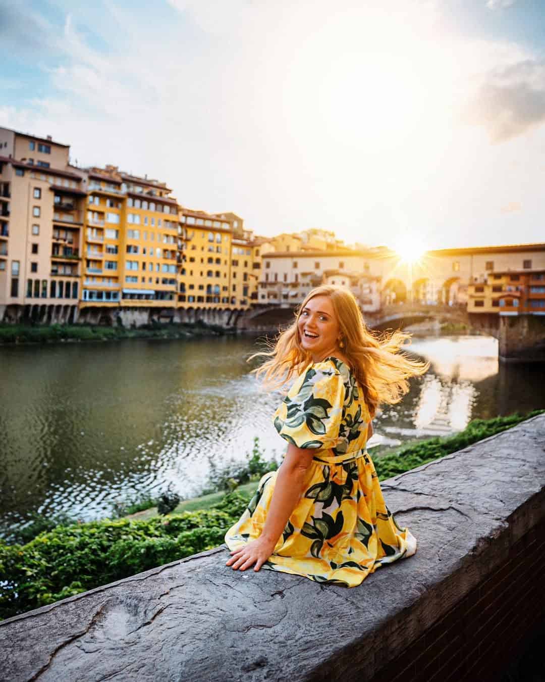 Smiling woman in a yellow dress sits on a stone ledge by a river at sunset, with buildings in the background&mdash;capturing the perfect moment for any Florence Italy itinerary.