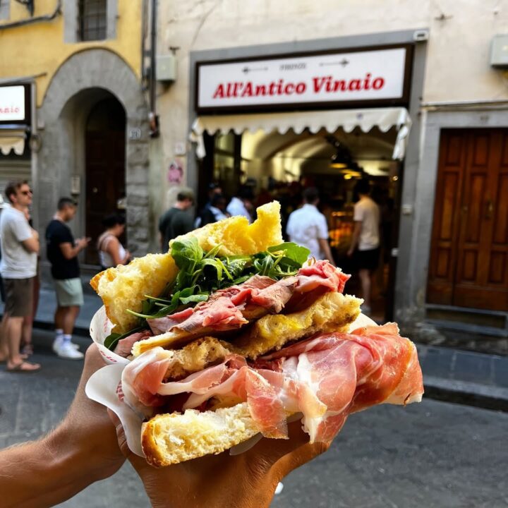 A hand holds a loaded sandwich with cured meats and greens outside All&rsquo;Antico Vinaio shop, capturing the essence of Florence food in the heart of the city.