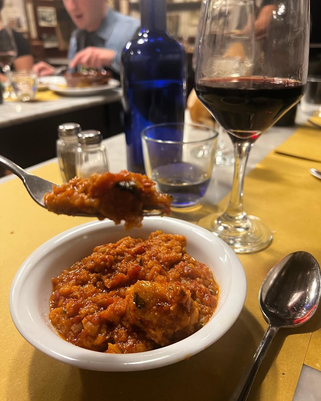 A bowl of chunky tomato stew, inspired by Florence food, sits on a yellow tablecloth with wine, water, and a spoonful being lifted.