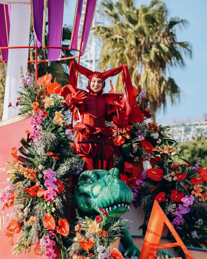 A person in a bright red costume stands on a float decorated with flowers and a green dragon, outdoors in sunlight&mdash;a vibrant scene often found at world festivals for those who love travel and unique celebrations.