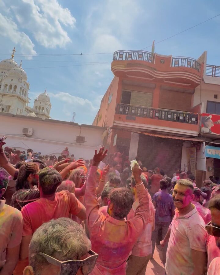 A crowd celebrates Holi with colorful powder outdoors, near a temple and orange building under a blue sky, making it one of the best festivals to experience during your travel.
