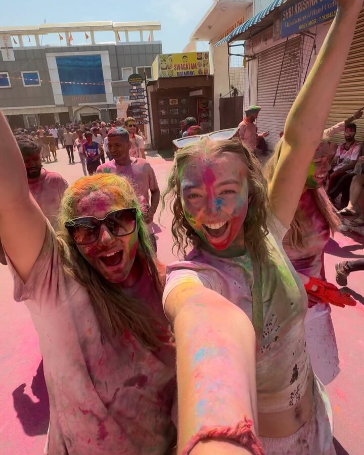 Two women covered in colorful powder celebrate Holi, smiling and raising their arms in a lively street scene&mdash;an unforgettable moment from one of the world&rsquo;s best festivals for those who love to travel.