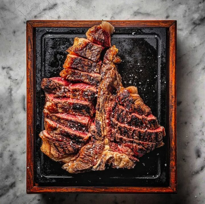 A sliced, grilled T-bone steak on a black tray with a wooden frame, set on a marble surface&mdash;a true homage to Florence food.