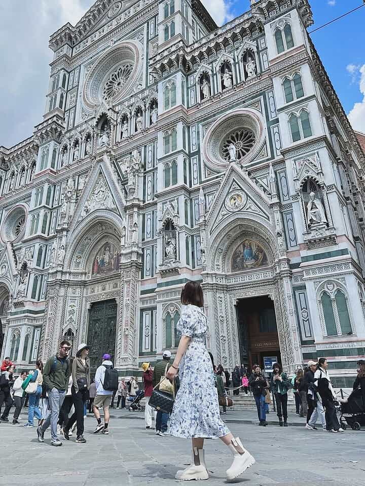 A woman in a floral dress stands in front of Florence Cathedral, a highlight on any Florence Italy itinerary, as tourists stroll nearby under a clear blue sky.