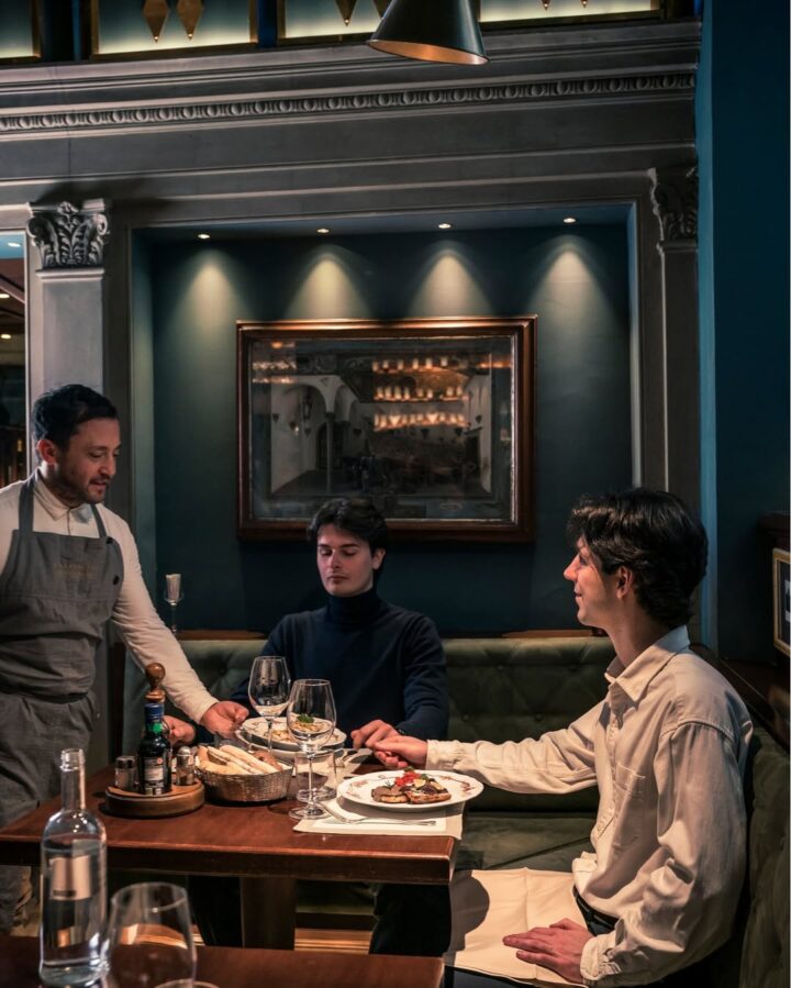 Two men hold hands at a restaurant table while a waiter serves them delicious Florence food and drinks in a cozy setting.