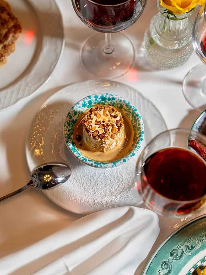 A small dessert topped with nuts sits on a plate beside a spoon and two glasses of red wine on a white tablecloth&mdash;perfect for savoring during your Florence Italy itinerary.