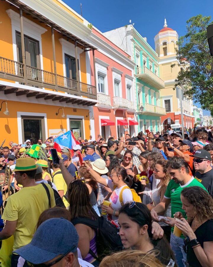 A crowd celebrates on a colorful street with a Puerto Rican flag, under a bright blue sky&mdash;capturing the vibrant spirit of travel and some of the best festivals in the Caribbean.