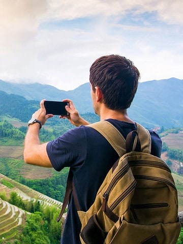 A man with a backpack takes a photo of green rice terraces and mountains with his phone.