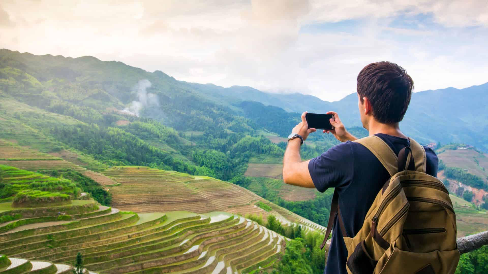 A man with a backpack takes a photo of green rice terraces and mountains with his phone.