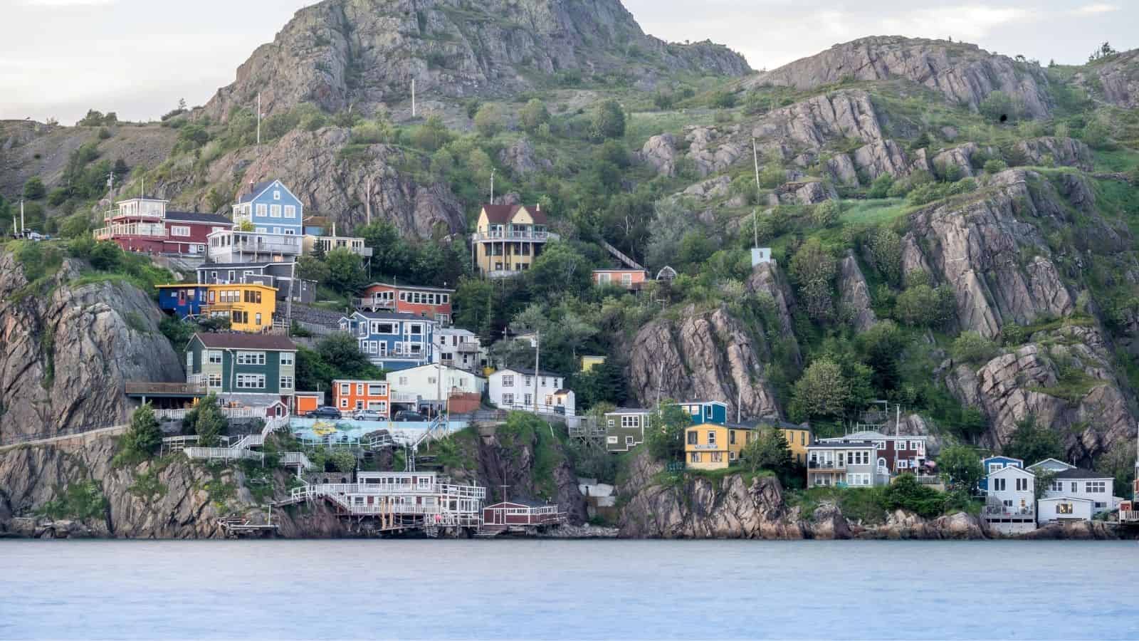 Colorful houses are built on a rocky hillside above the water, surrounded by greenery and cliffs.