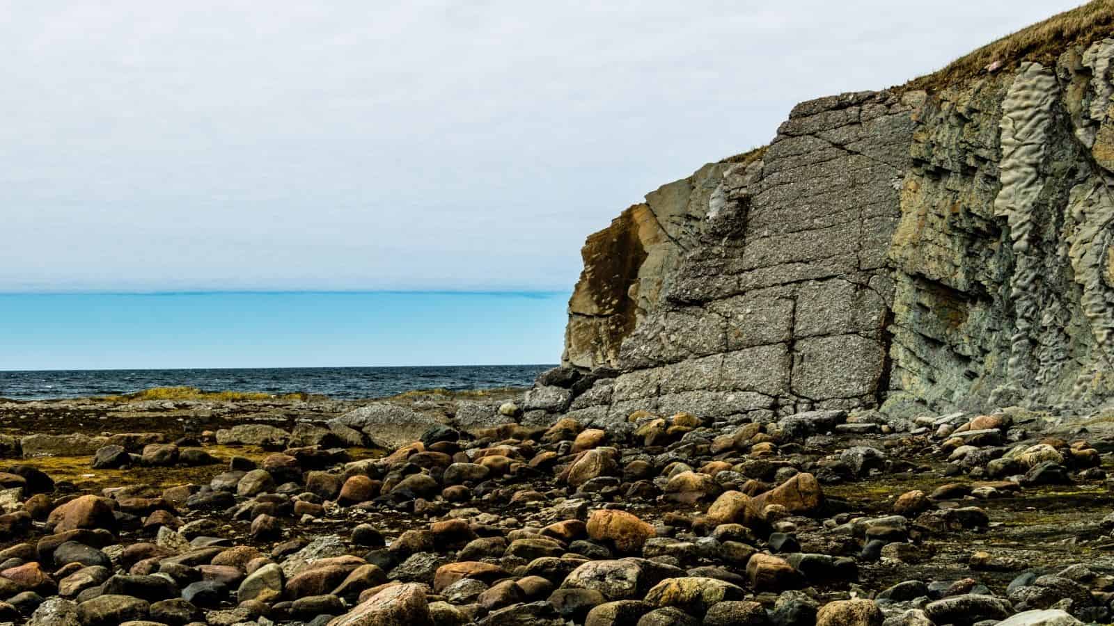 Rocky shoreline with a large stone cliff beside a calm sea under a partly cloudy sky&mdash;perfect for those seeking scenic Canada travel and unforgettable Canadian vacations.