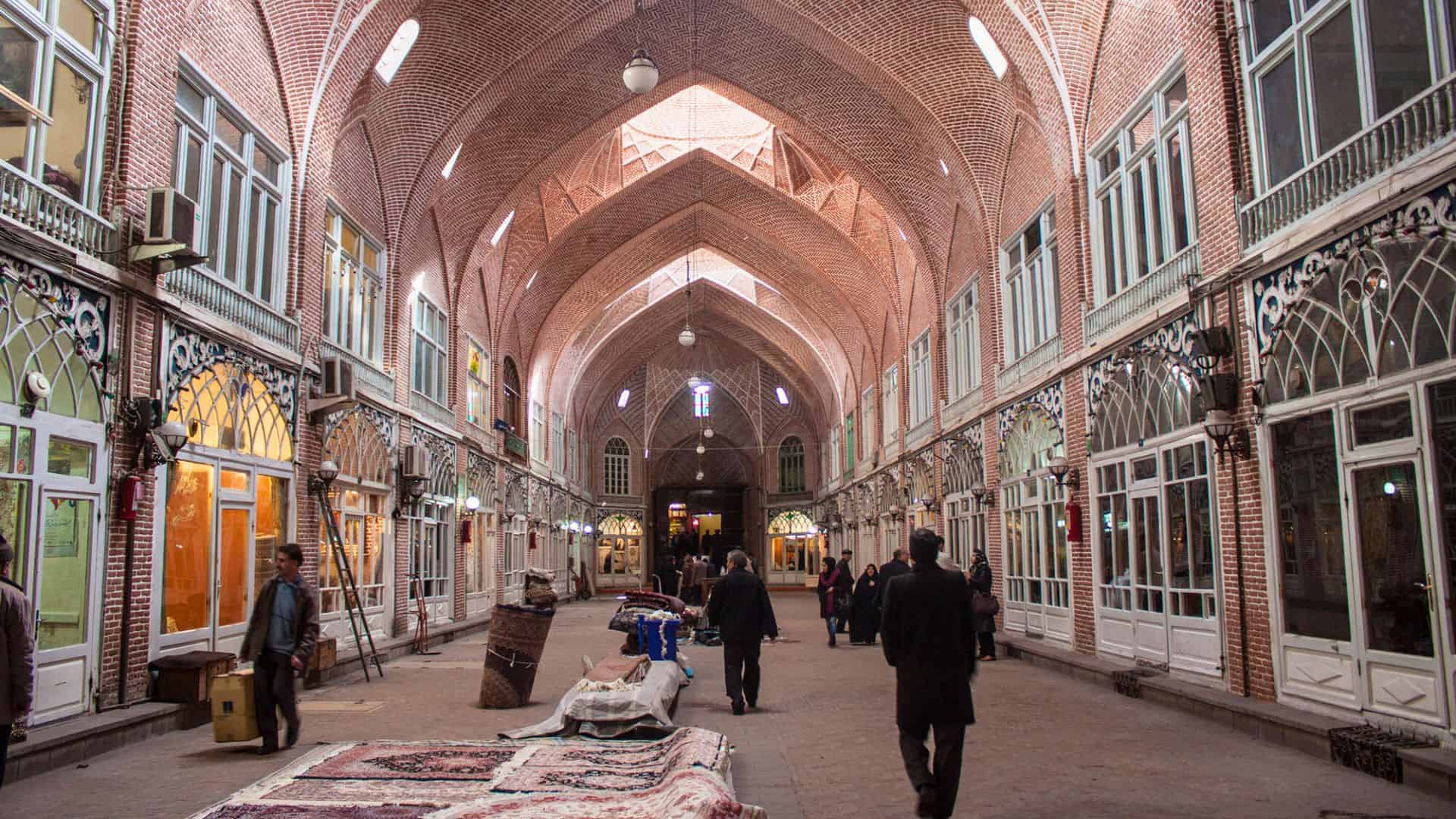 People walk through a covered market hall with brick arches, large windows, and carpets on display.
