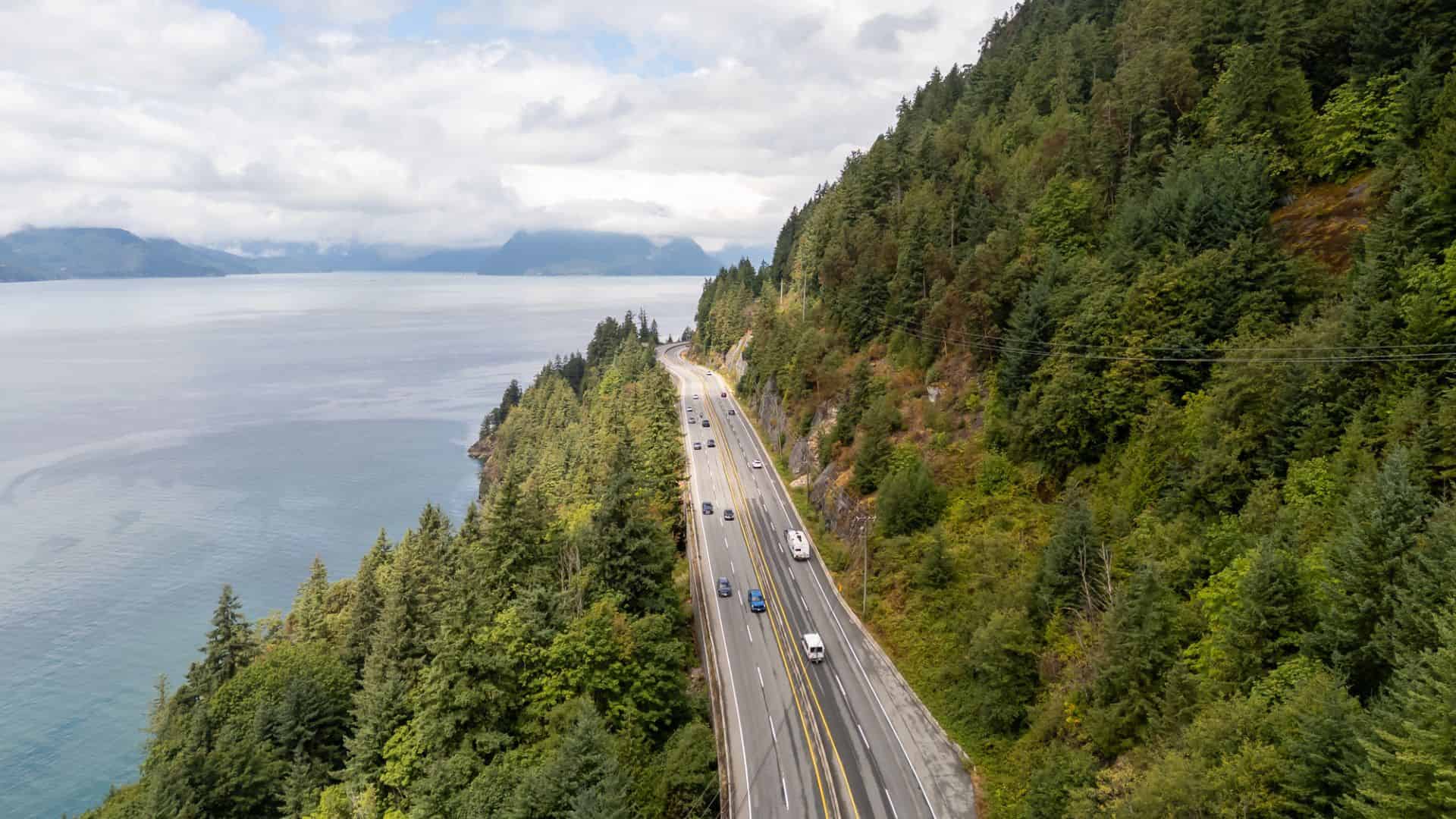 A coastal highway runs between a forested hillside and the ocean, with cars driving in both directions.