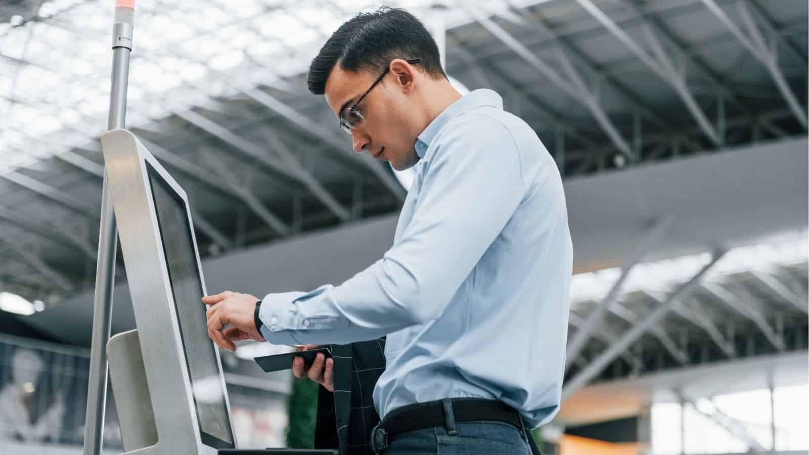 A man uses a self-service kiosk at the airport, holding his passport and checking the screen to avoid boarding line mistakes and steer clear of annoying passengers.