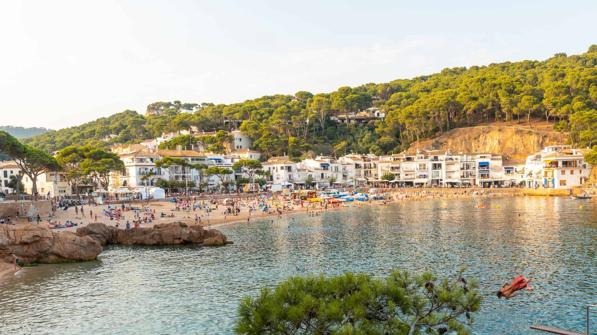 A crowded beach with clear water, white buildings, and green hills in the background on a sunny day.