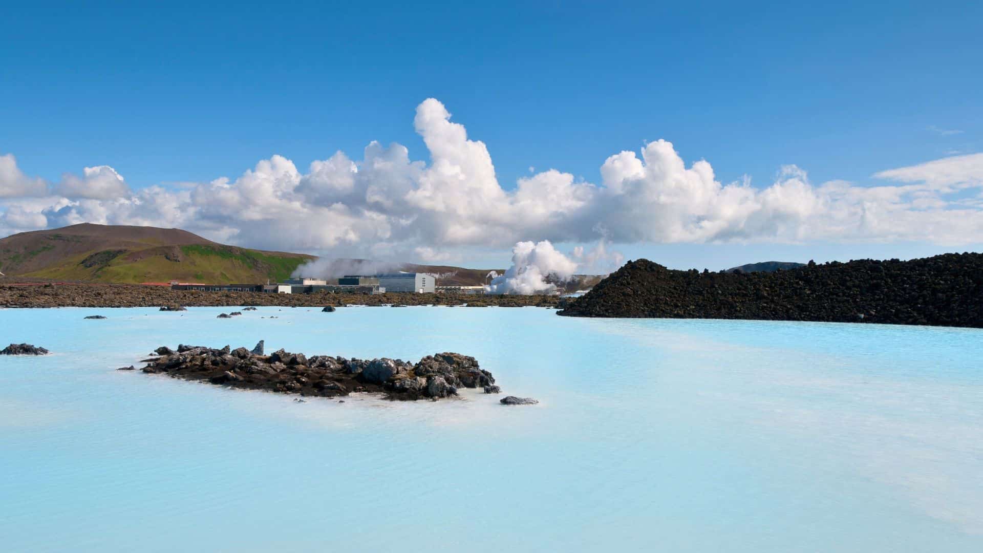 Milky blue geothermal lagoon with rocky islands, steam rising in the distance, and a bright blue sky with clouds.
