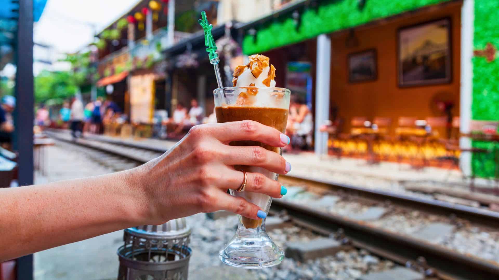 A hand holding an iced coffee by a railway track with cafes and colorful buildings in the background.