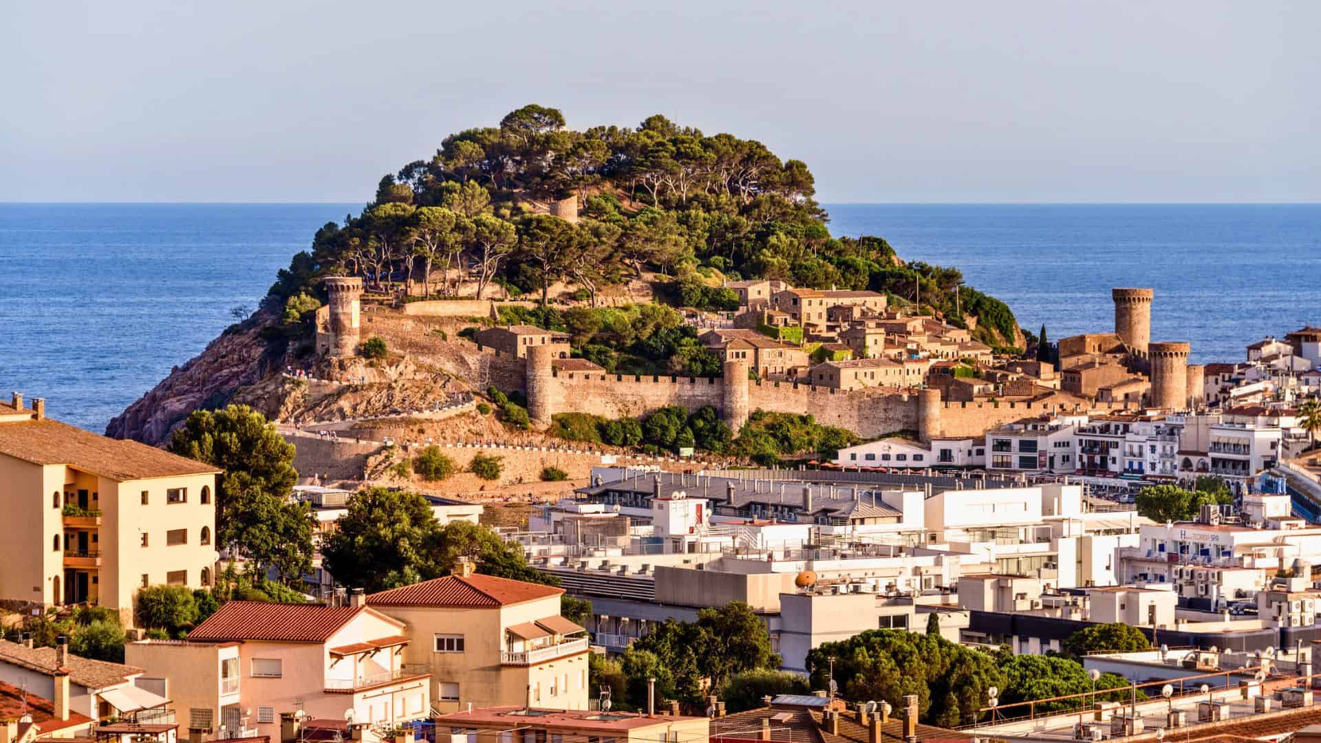 Medieval castle with stone walls on a hill by the sea, overlooking a coastal town with white buildings.