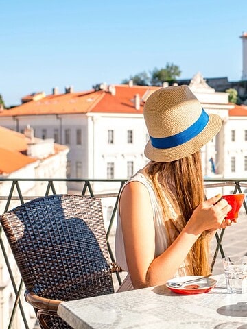 Woman in a hat drinks coffee at a balcony café overlooking a sunny European town square with historic buildings.