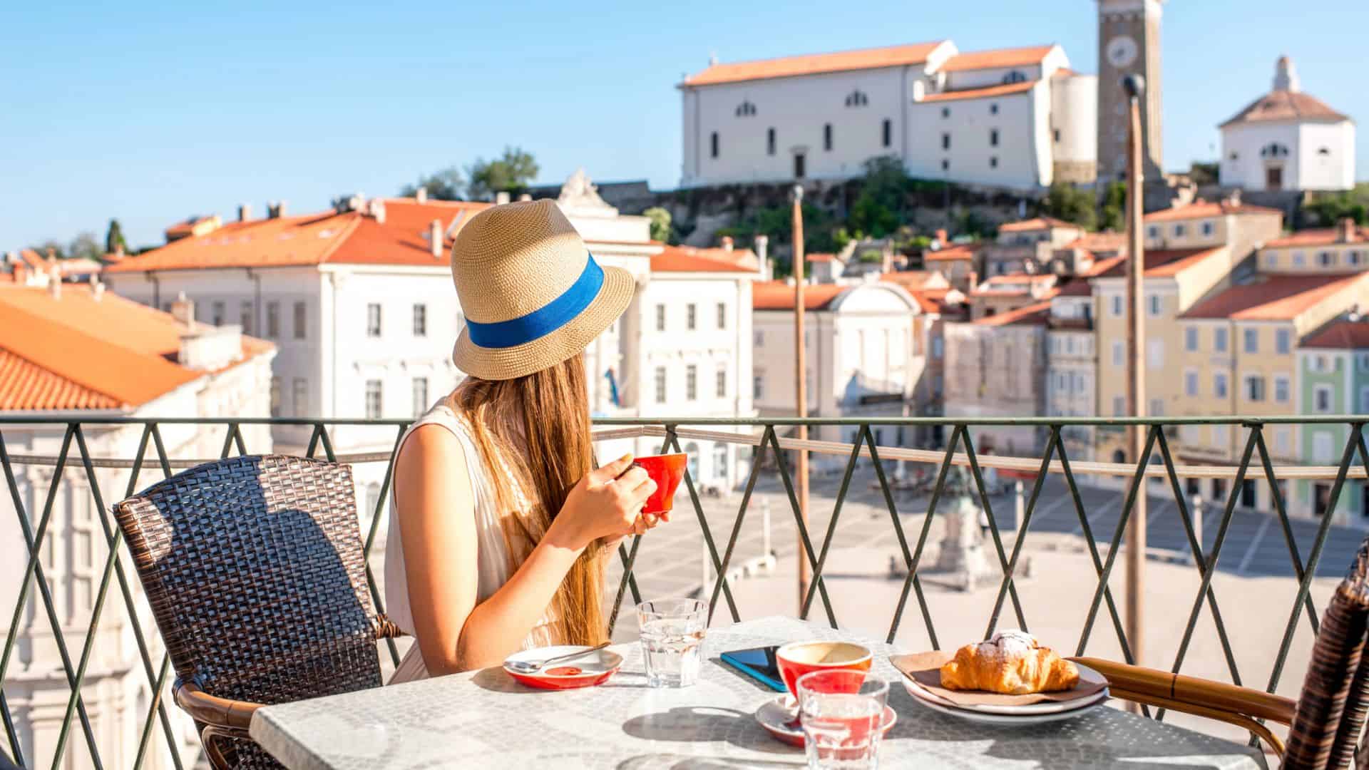 Woman in a hat drinks coffee at a balcony caf&eacute; overlooking a sunny European town square with historic buildings.