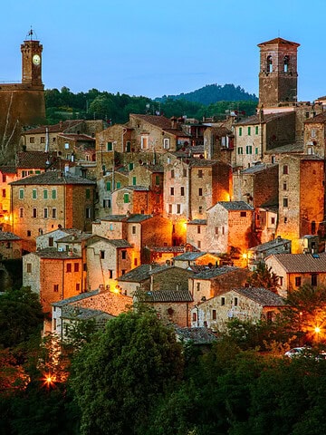 Hilltop Tuscan village at dusk, with old stone buildings and warm streetlights glowing among the houses.