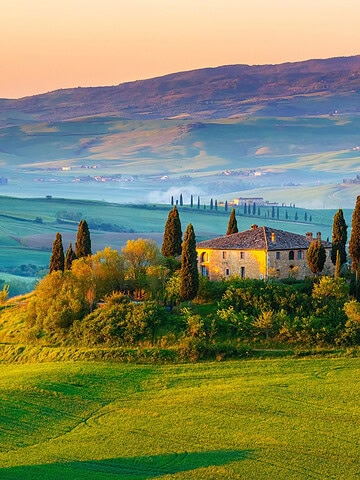 Stone farmhouse surrounded by trees on a green hill, with mountains in the background at sunset—a perfect scene for any Tuscany travel guide.