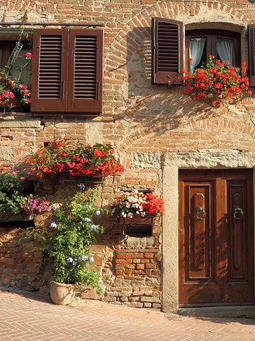 Wooden door and shuttered windows on a brick wall, reminiscent of charming Tuscany villages, decorated with vibrant potted flowers and plants.