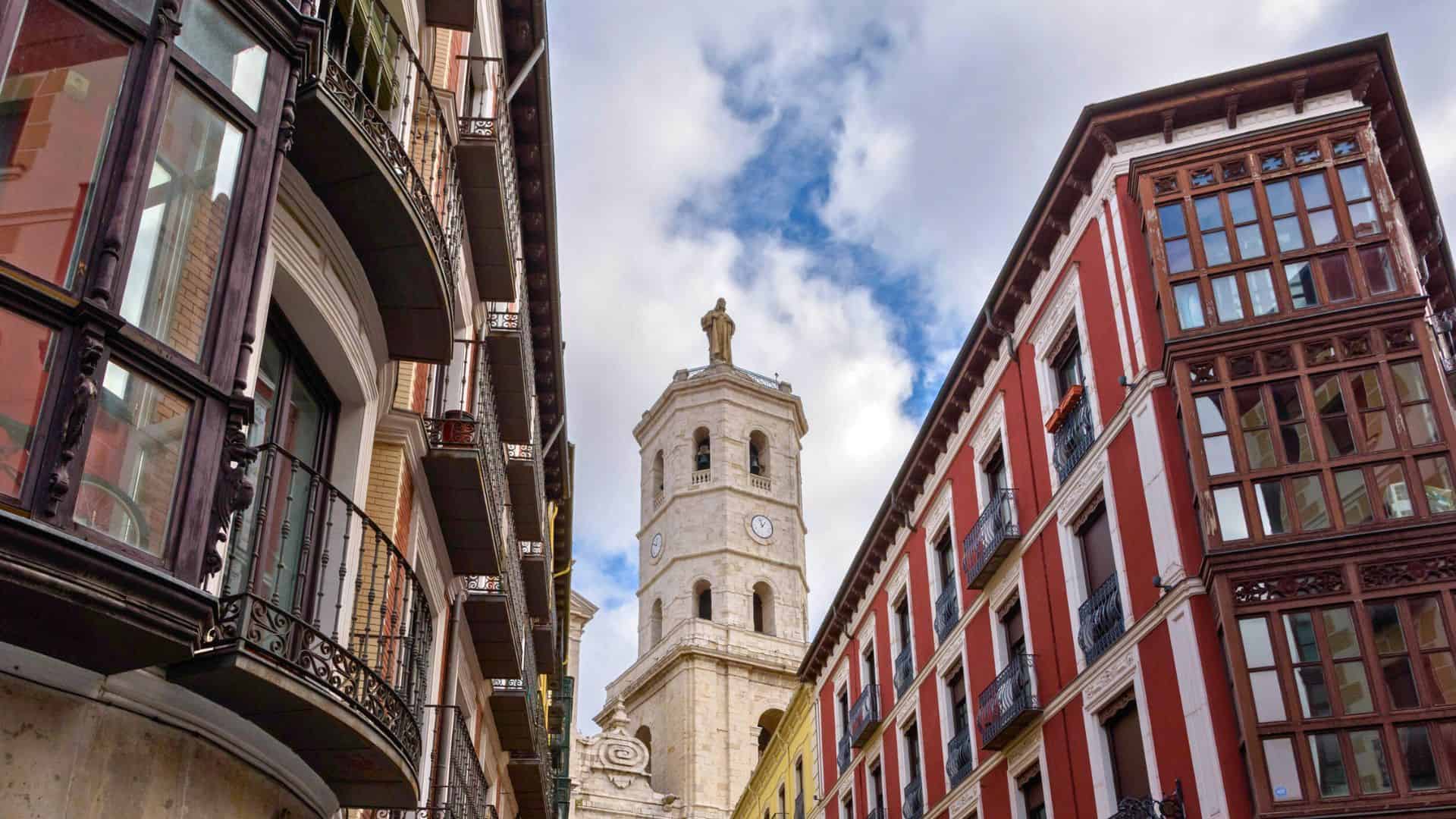 View of a stone clock tower between two buildings with balconies under a partly cloudy sky.