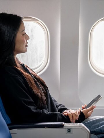 Woman sitting in an airplane seat by the window, holding a smartphone and looking outside.