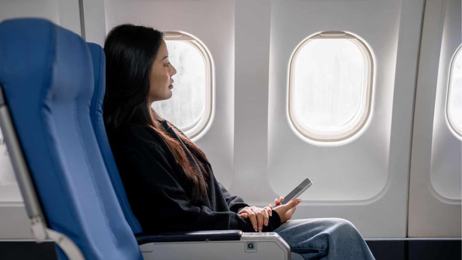Woman sitting in an airplane seat by the window, holding a phone and looking outside.